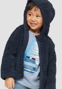 Navyblauwe donzige zip-up hoodie met een gestructureerde afwerking, voorzien van een capuchon. Draag het over een lichtblauw shirt met een afbeelding van een politieauto.