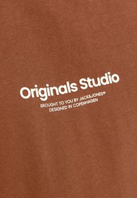 Tela de algodón marrón con texto blanco: "Originals Studio," "Traído a ti por JACK & JONES," y "Diseñado en Copenhague."