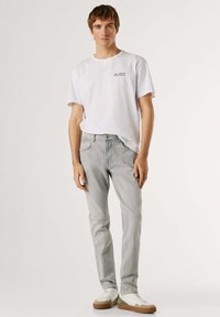 Joven de pie, vistiendo una camiseta blanca con estampado, jeans gris claro y zapatillas blancas con suelas marrones sobre un fondo liso y claro.