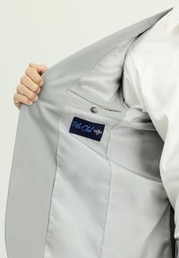 Innenseite eines hellgrauen Sakko mit glatter Fütterung, ausgestattet mit einem blauen Etikett, auf dem "Polo Club" steht, und einer Tasche mit Knopfverschluss.