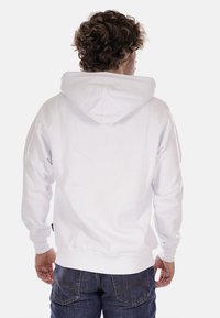 Felpa bianca in morbido materiale, con tasca a marsupio, vestibilità comoda e cappuccio con cordino. Indossata con jeans scuri.