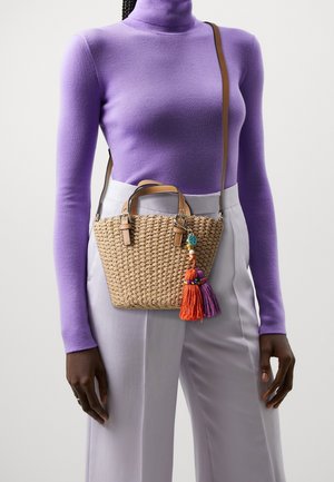 Osoba oblečená v fialovém roláku a světlých kalhotách, nesoucí pletenou slaměnou kabelku s koženými pásky a barevnými střapcovými přívěsky na klíče.