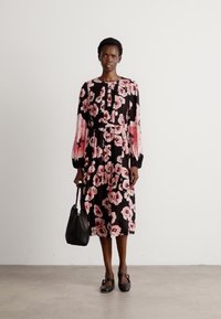 Vestido negro con estampado floral en rosa, de mangas largas y cuello alto. Combinado con un bolso negro y zapatos planos negros. Textura de tela suave.