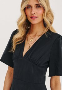 Černé šaty s hlubokým V výstřihem, krátkými puff rukávy a nařasenými detaily na prsou. Zlaté vrstvené náhrdelníky podtrhují outfit.