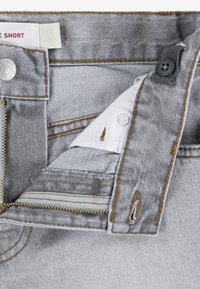 Γκρι τζιν σορτς με φερμουάρ μπροστά και υλικά σε ρευστό χαλκού, αντίθετη ραφή και λευκή υφασμάτινη επένδυση στο εσωτερικό της μέσης.