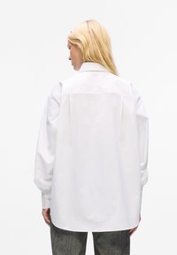 White oversized button-up shirt with a classic collar, long sleeves, and a smooth texture. Features a subtle back yoke detail.