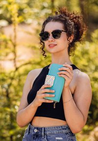 Turquoise wallet with a white snap closure featuring a colorful polka dot design. Smooth texture with a zipper detail and brand name visible.