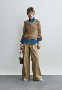 Jeune personne portant un pull en maille torsadée beige sur une chemise en denim bleu, pantalon beige à jambes larges, chaussures blanches, tenant un petit sac à main marron.