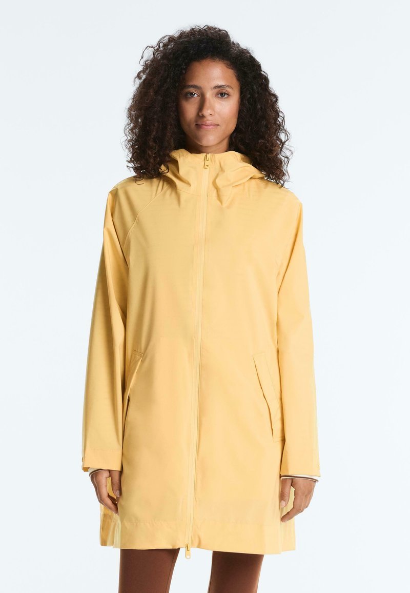Femme aux cheveux bouclés portant un long imperméable jaune à capuche avec des poches avant et une fermeture éclair, debout devant un fond uni.