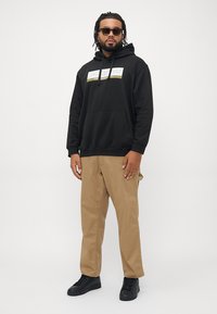 Man with sunglasses wearing black hoodie with front logo, khaki pants, and black sneakers standing against plain light background.