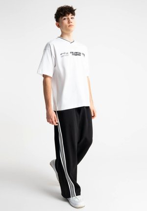 Młoda osoba stoi, ubrana w białą koszulkę z napisem "Following Forward", czarne spodnie z białymi paskami oraz białe trampki na białym tle.