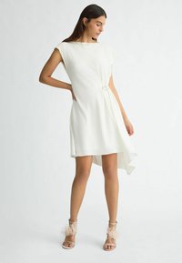 Abito bianco asimmetrico con maniche a cappuccio, caratterizzato da un dettaglio arricciato laterale con un accento circolare. La modella indossa dei sandali con tacco rosa pallido e piume.