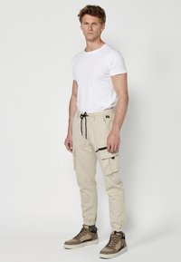 Beige Cargo-Hosen mit Reißverschlusstaschen, elastischem Bund mit Kordelzug, schlankem Schnitt, kombiniert mit braunen und weißen Sneakers.