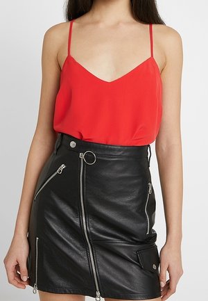 Vrouw draagt een rood spaghettihemd dat in een zwarte leren minirok is gestopt, met meerdere zilveren ritsen en een ringdetail.