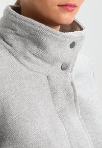 Mujer vestida con un abrigo de lana gris claro de cuello alto, con dos botones de presión plateados visibles cerca del cuello.