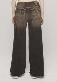 Hnědé džíny s širokými nohavicemi a vybledlým vzhledem, kontrastním šitím, zadními kapsami s dekoračními prvky a nášivkou na pase.