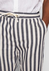 Persoon in beige en marineblauwe verticaal gestreepte broek met trekkoord en een wit shirt, hand in de zak.