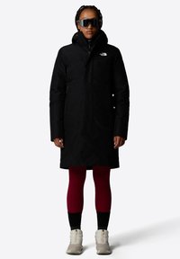 Zwarte geïsoleerde parka met een hoge kraag en ritssluiting, voorzien van een klein wit logo. Model draagt rode leggings en crèmekleurige laarzen.