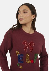 Pull bordeaux avec un motif festif comprenant un nœud rouge à paillettes, les lettres "E.L.F." multicolores à paillettes, et des accents d'étoiles dorées.
