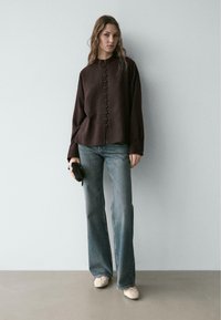 Dunkelbraunes Hemd mit Knopfleiste und hohem Kragen, kombiniert mit ausgewaschenen blauen Schlagjeans und hellbeigen flachen Schuhen. Einfaches schwarzes Clutch-Accessoire.