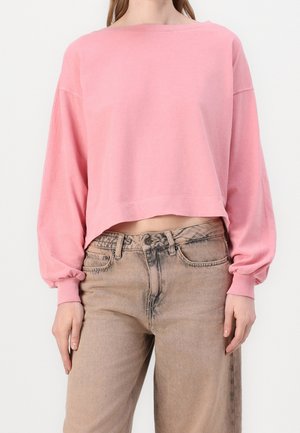 Kobieta ubrana w luźną, różową, skróconą bluzę z długimi rękawami oraz wysokim stanem, wyblakłe brązowe dżinsy na tle białego tła.