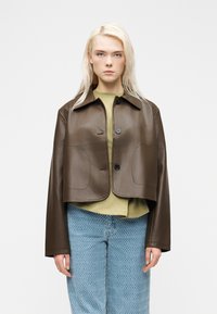 Young person with long blond hair wearing a cropped brown leather jacket over a green shirt and patterned blue jeans, standing against white background.