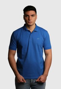 Polo blu in cotone, con colletto a due bottoni, maniche corte e un piccolo logo sul lato sinistro del petto. Tessuto testurizzato.