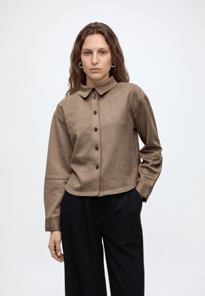 Femme aux longs cheveux bruns ondulés porte une chemise beige boutonnée et un pantalon noir, debout devant un fond gris clair uni.