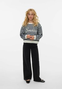 Pull rayé bleu marine et blanc avec poignets côtelés, associé à un pantalon noir ample et des chaussures plates noires. Le mannequin est debout sur un fond blanc.