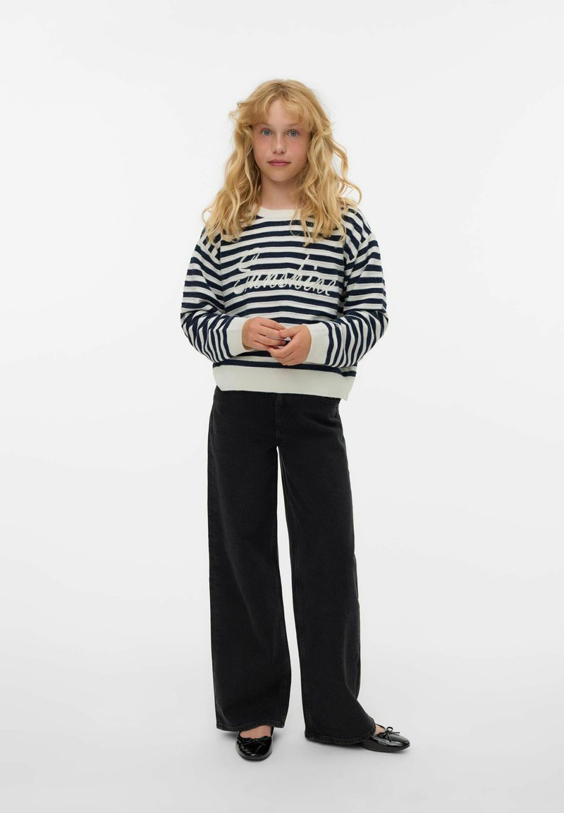 Pull rayé bleu marine et blanc avec poignets côtelés, associé à un pantalon noir ample et des chaussures plates noires. Le mannequin est debout sur un fond blanc.