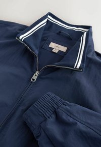 Veste zippée bleu marine avec poignets élastiques, col côtelé avec des rayures blanches et étiquette de marque "NEXT" à l'intérieur du col.