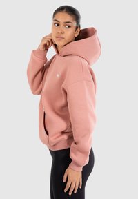 Sweatshirt à capuche rose avec une coupe décontractée, grande poche avant, poignets côtelés et un petit logo sur la poitrine. Tissu doux et texturé.