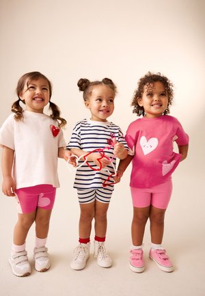Tre bambini piccoli sorridenti stanno in piedi tenendosi per mano; due indossano completi con cuori rosa, e uno indossa una maglietta a righe e pantaloncini, tenendo in mano delle fil di ferro a forma di cuore.
