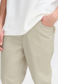 Pantalones color beige claro de algodón, con un corte relajado y bolsillos laterales. La tela tiene una textura suave con detalles de costura sutiles.