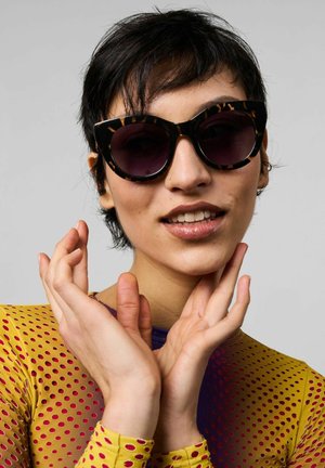 Gafas de sol de ojo de gato con una montura de carey y lentes oscuros. El modelo lleva un body de malla colorido y con puntos. Manos cerca de la cara.