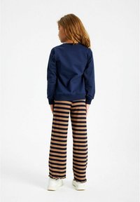 Bambino con capelli lunghi che indossa una felpa blu navy, pantaloni a strisce marroni e blu navy, e scarpe bianche, in piedi con la schiena rivolta verso la fotocamera su uno sfondo bianco.