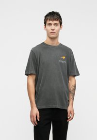 Graues, übergroßes T-Shirt mit Rundhalsausschnitt und kurzen Ärmeln. Die Vorderseite zeigt orangefarbene, grafische Schriftzüge. Wird mit schwarzen Hosen getragen.