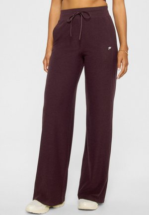 Woman wearing loose maroon sweatpants with drawstring waist and white sneakers, standing against plain background.