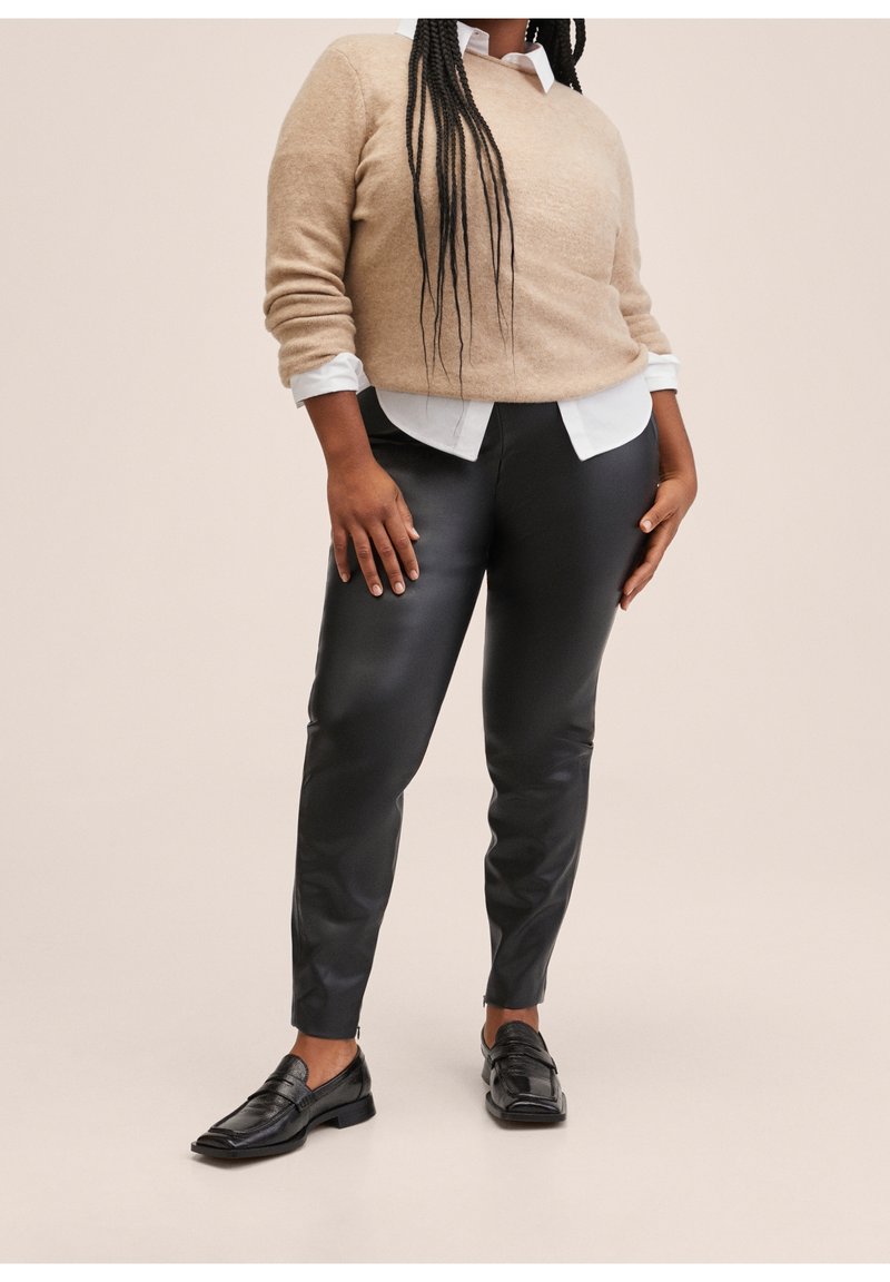 Světlý béžový svetr, pod ním bílá košile s límečkem, lesklé černé kalhoty z umělé kůže a černé mokasíny. Hladká textura se jemnými akcenty.