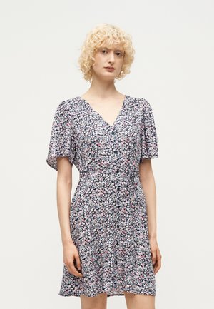 Femme aux cheveux blonds bouclés portant une robe courte à manches avec des boutons sur le devant, à motif floral, col en V et ceinture nouée à la taille, debout devant un fond uni.