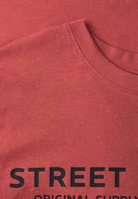 T-shirt en coton rouge avec col rond et manches courtes. "STREET ORIGINAL SUPPLY" est imprimé en lettres noires épaisses sur le devant.