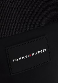 Schwarzer Stoff mit einem genähten Aufnäher, der das "Tommy Hilfiger"-Logo und ein rot-weiß-blaues Flaggen-Symbol auf einer Tasche oder einem Bekleidungsstück zeigt.