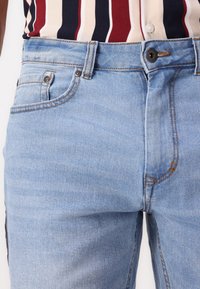 Close-up of light blue denim jeans with visible button, zipper, pockets, and brown stitching, paired with a vertically striped shirt.