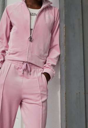 Personne portant un survêtement en velours rose clair avec la veste fermée à moitié et un pantalon à cordon de serrage, debout contre un mur gris et blanc.