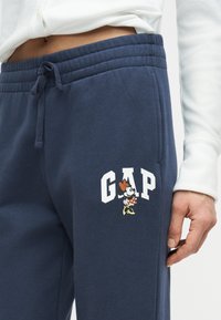 Σκούρο μπλε φόρμας με κορδόνι στη μέση, διακοσμημένη με λευκό κείμενο "GAP" και ένα μικρό γραφικό της Μίννι Μάους στο μηρό, φορεμένη με λευκό τοπ.