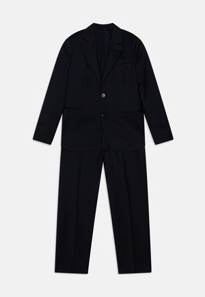 Musta puku, joka koostuu räätälöidystä takista, jossa on kaksi nappia ja yhteensopivat housut. Asussa on hienovarainen raidoituskuvio ja teksturoitu vuori.