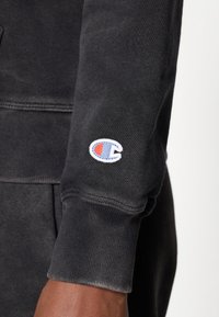 Musternööpidega must dressipluus, mille varrukatel on väike brodeeritud mitmevärviline logo. Pehme tekstuur ja lõdva disain.