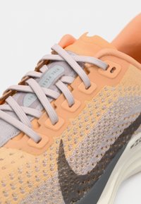 Prim-plan al unei adidași tricotate în nuanțe de piersică și gri, cu șireturi gri, logo-ul Swoosh negru și talpă amortizată.