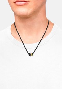 Collana intrecciata nera con tre perline cilindriche: due nere e una gialla, indossata su una semplice camicia bianca.