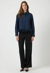 Vrouw die staat in een donkerblauwe blouse met knopen en een gerimpelde zoom, zwarte wijde broek en zwarte puntige hakken tegen een witte achtergrond.
